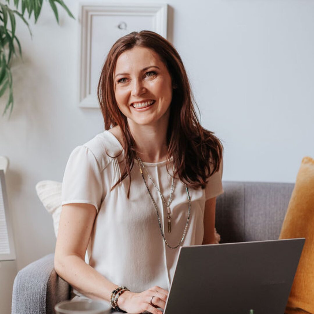 brunette woman smiling sitting on a grey couch with a laptop.